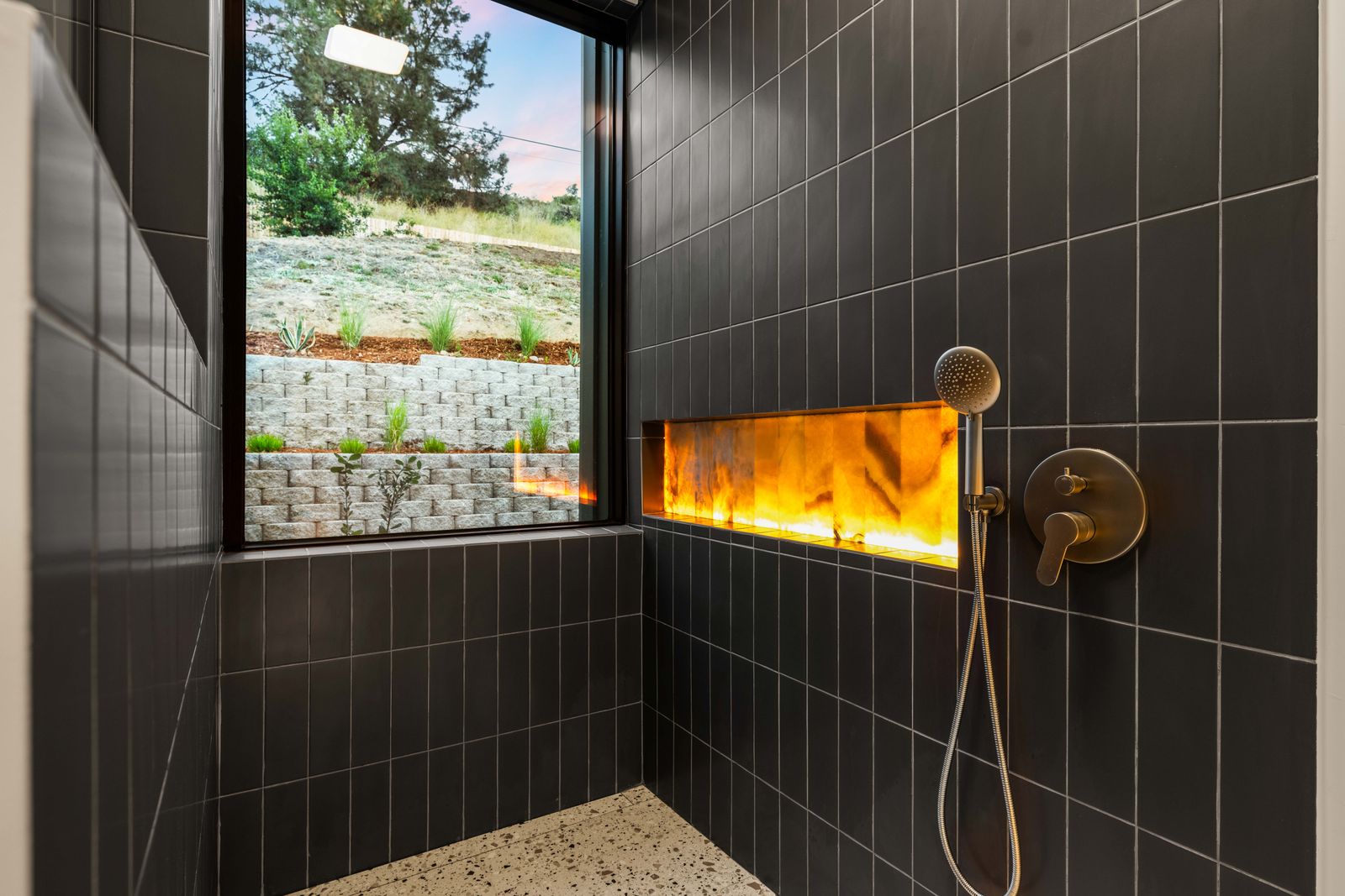 Walk-in shower with black tile, brass fixtures and backlit onyx niche